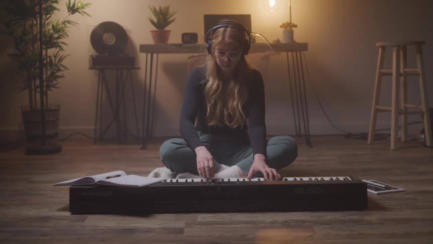 A young woman with long reddish brown hair wearing headphones and glasses sits cross legged on a wooden floor playing an electric keyboard in a dimly lit room with a warm cozy ambiance