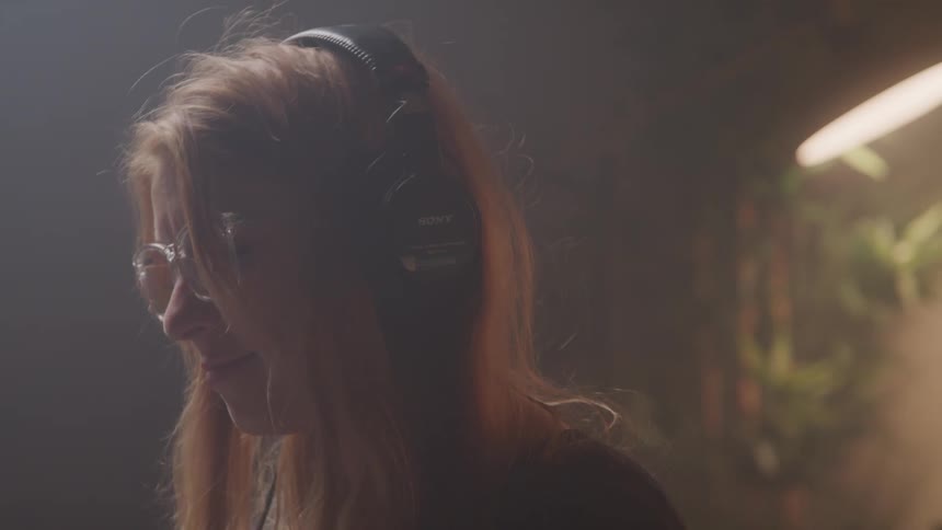 A woman with reddish hair and glasses bobs her head to music while wearing sony headphones in a softly lit room with plants glowing in the background