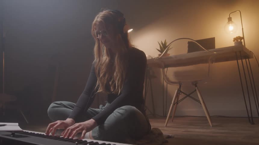 A young woman with long hair and headphones sits cross legged on the floor playing a keyboard in a dimly lit room with a desk and lamp