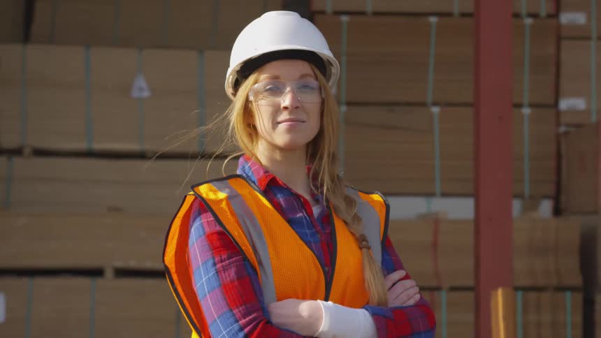 Female worker standing near a large amount of stacked lumber during the daytime smiles slightly