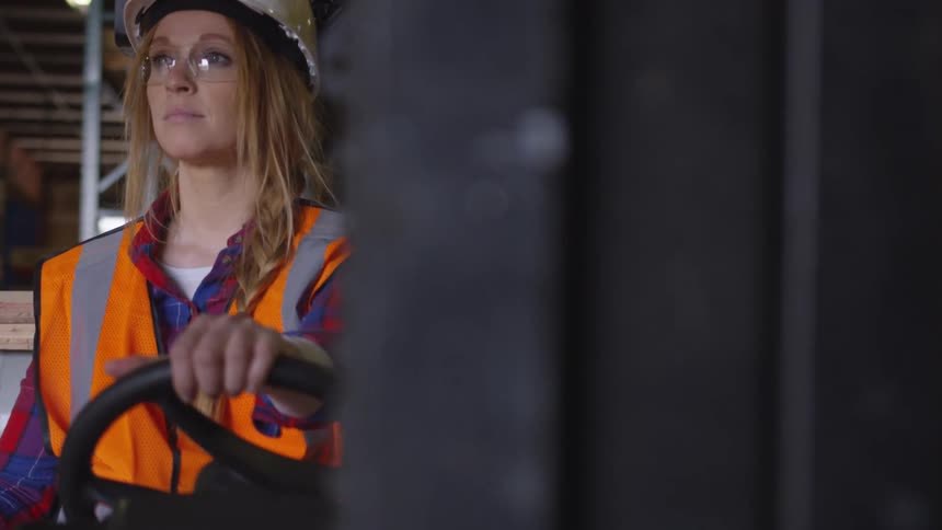 A woman in safety gear sits stationary inside a forklift as the camera slowly pans past the forklift fork to reveal her in a warehouse setting