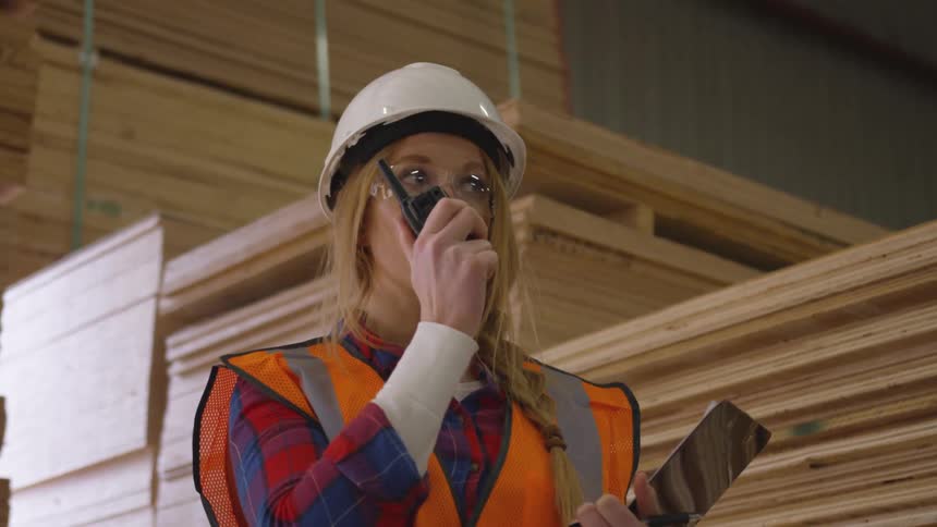 Woman in a hardhat holds a clipboard and talks into a walkie talkie