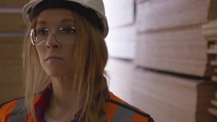 Woman standing in a warehouse filled with wood staring straight ahead without blinking