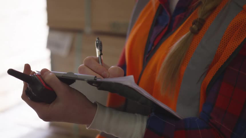 A woman is writing on her clipboard in the warehouse with a pen