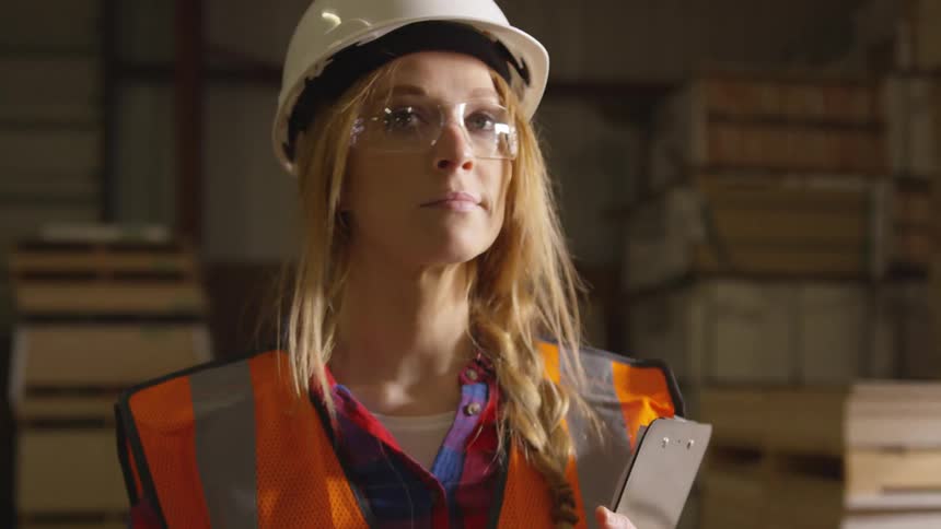 Woman in a hardhat holds a clipboard while walking through a warehouse