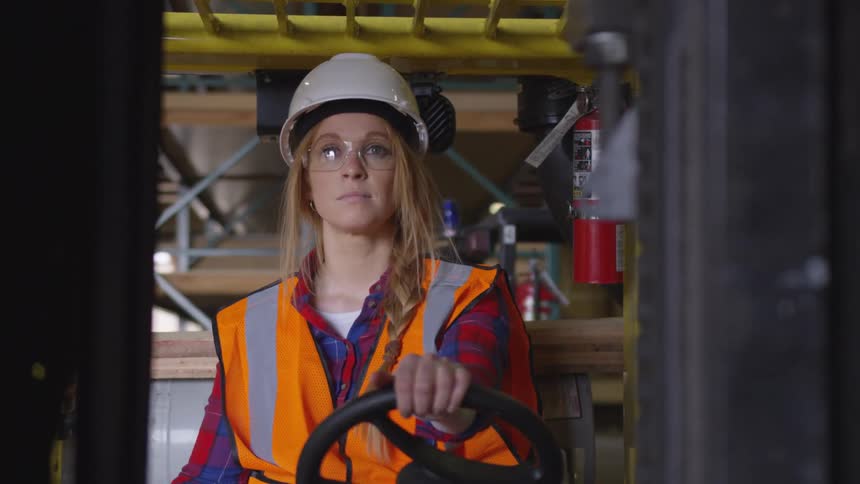 A blonde woman is driving heavy equipment at a construction site wearing a white hard hat safety glasses and an orange hazard vest over a long sleeve plaid shirt