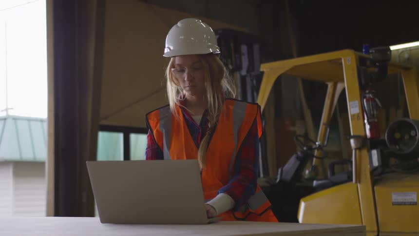 A worker wearing a hardhat and safety glasses is looking at a laptop screen in a garage