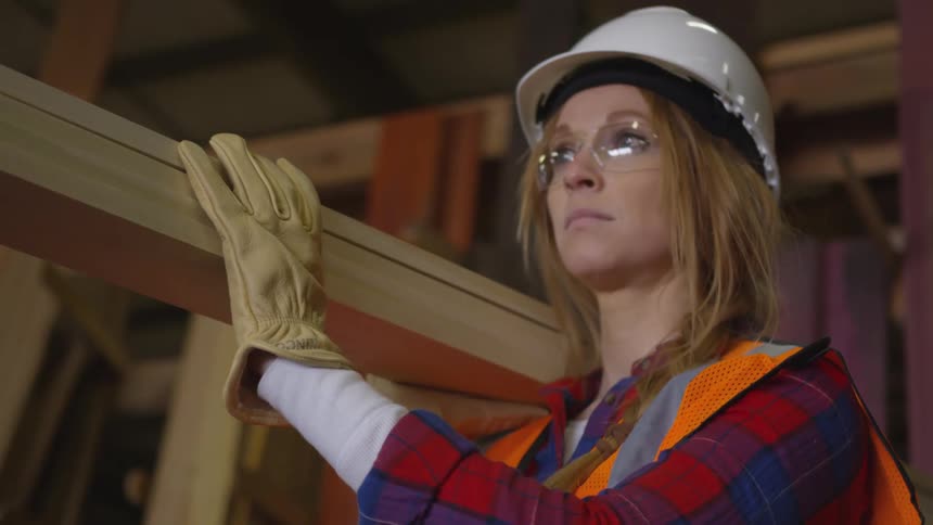 A woman wearing safety gear holds wooden planks on her shoulder inside a lumber facility focused and prepared for work