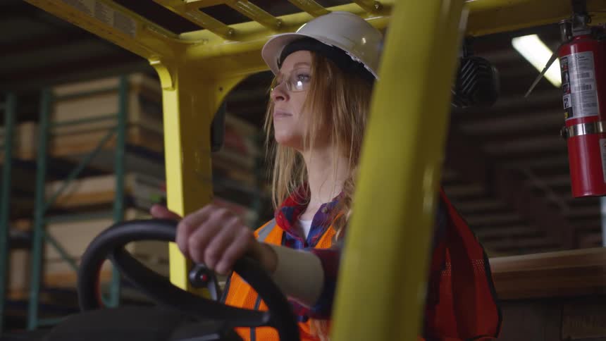 Woman rests her hand on the steering wheel of a forklift while sitting in it in a warehouse