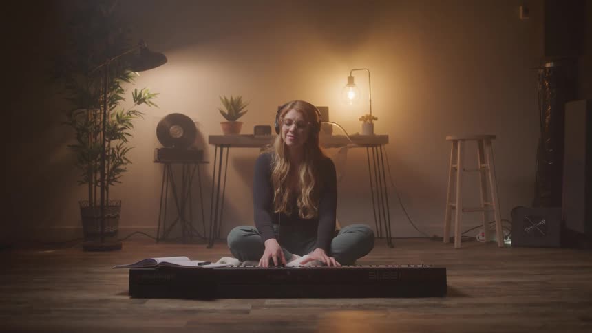 A young woman with long reddish brown hair wearing glasses and headphones sits cross legged on a wooden floor playing an electric keyboard in a dimly lit cozy room