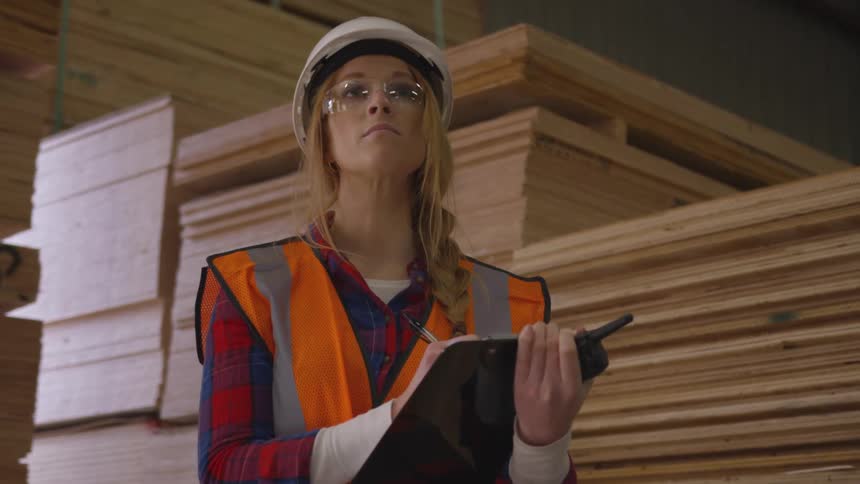 A woman wearing a safety vest and hard hat holds a clipboard in a lumberyard