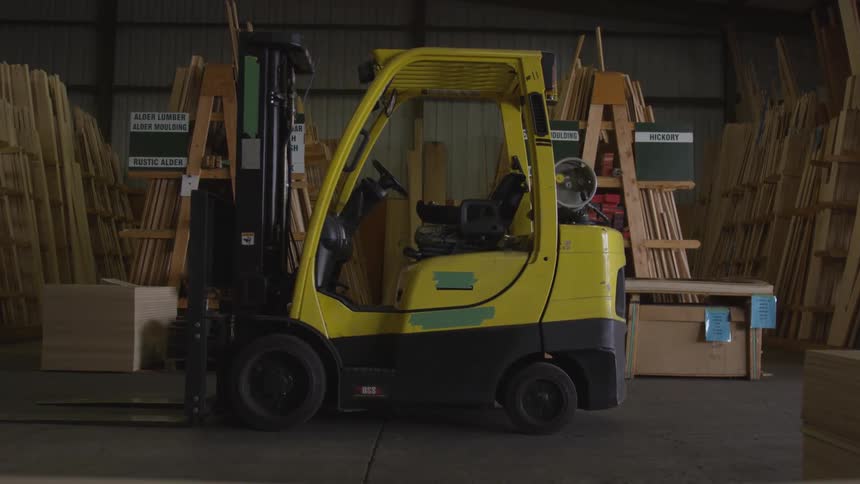 A yellow forklift sits idle in a lumberfilled woodshop bay surrounded by neatly organized wooden boards and materials