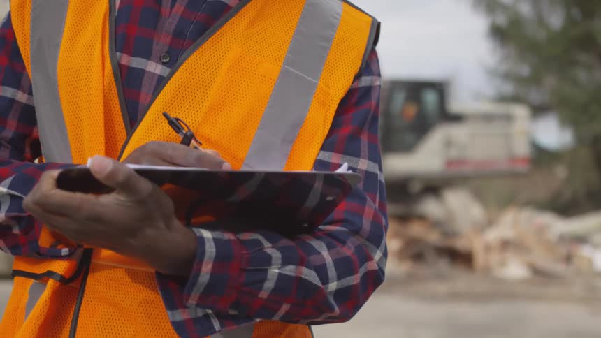 Man in a safety vest writes on a clipboard with a pen