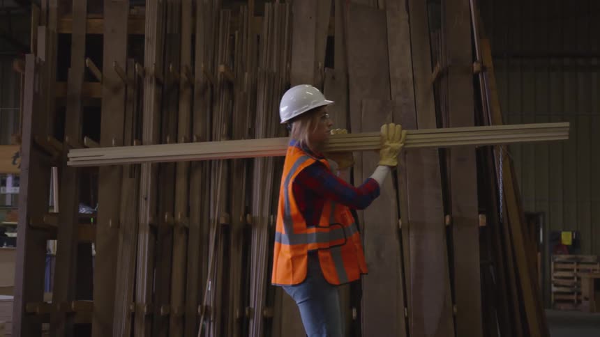 Woman carries planks of wood on her shoulder in a warehouse