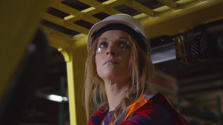 A woman wearing a hard hat safety glasses and work attire looks up in a warehouse