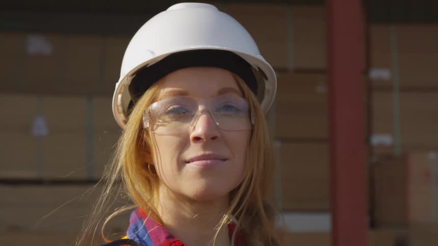 The lady in the safety glasses and the white hard hat turns to look at the camera while she is standing in front of a bunch of card board boxes and a square pole