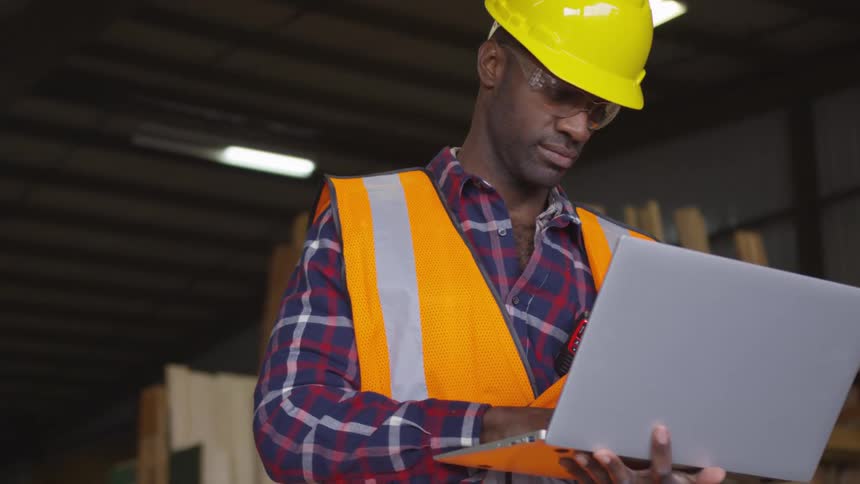 Construction worker standing in a room with lumber holds a laptop computer with one hand and types with the other hand