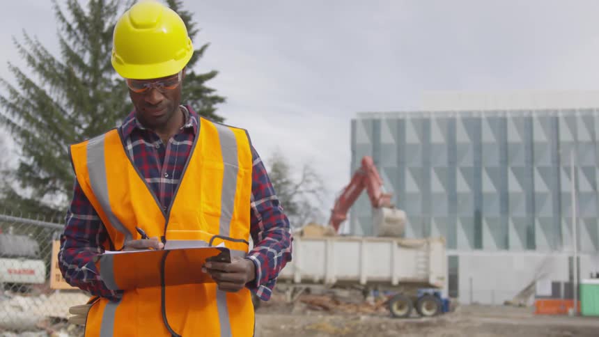 An employee is writing on a clipboard as the excavator is working