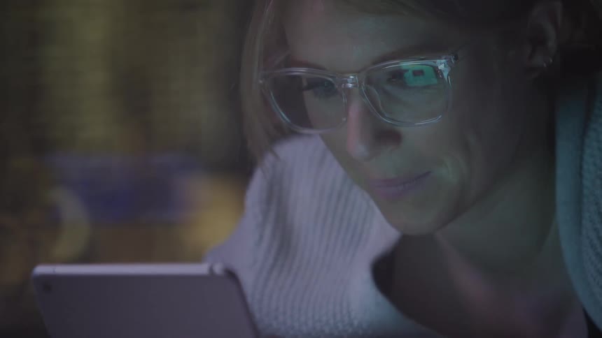 Woman smiles and scrolls on a tablet while her glasses reflect its screen