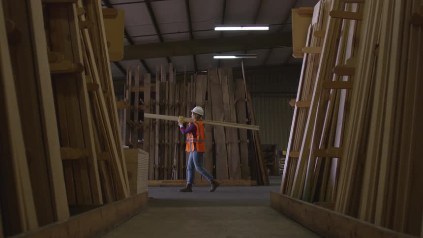 Woman supports lumber with both hands as she balances it on her shoulder to carry it through the warehouse