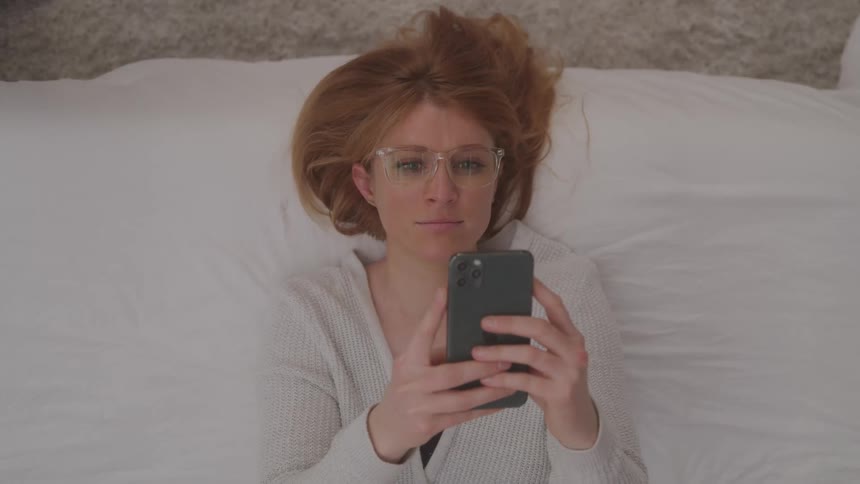 Woman with red hair laying down on bed scrolling on a cell phone