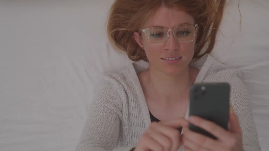Woman lying on her bed smiles while scrolling on her smartphone