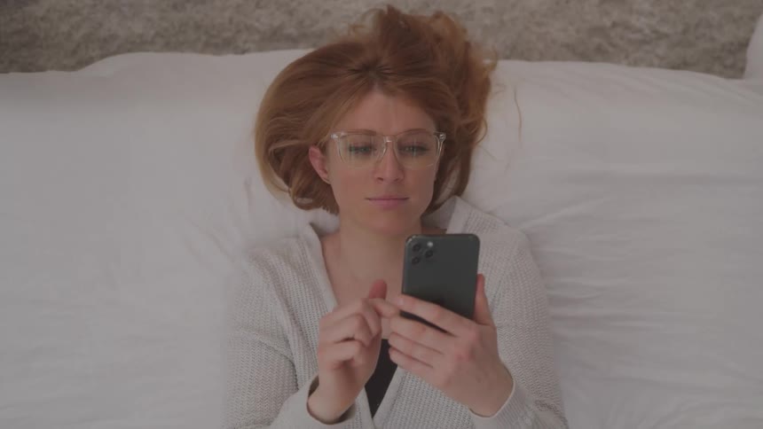 Woman scrolls on her phone while lying down in bed