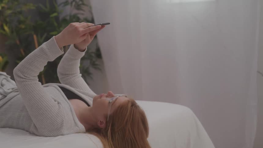 Redheaded woman lying on her back on her bed texts on her smartphone