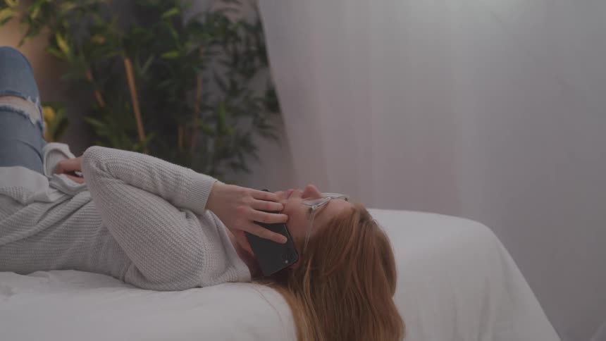 Woman with red hair laying on edge of bed talking on cell phone