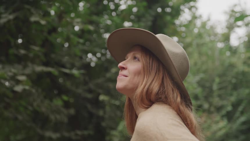 A woman with long light brown hair wearing a hat looks upwards in a hazelnut orchard with tall leafy trees