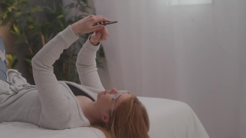 A woman with reddish brown hair wearing glasses and a light colored top lies on her back on a white bed holding a cell phone above her