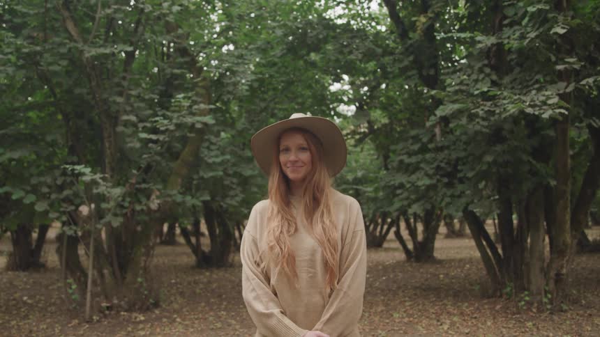 A young woman with long light brown hair and a wide brimmed hat smiles directly at the camera while standing in a hazelnut orchard surrounded by tall leafy trees