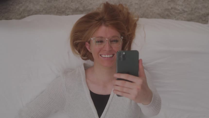 A smiling woman with light brown hair and glasses lies on her back on a white sheetcovered bed looking directly at the camera while holding a dark green iphone