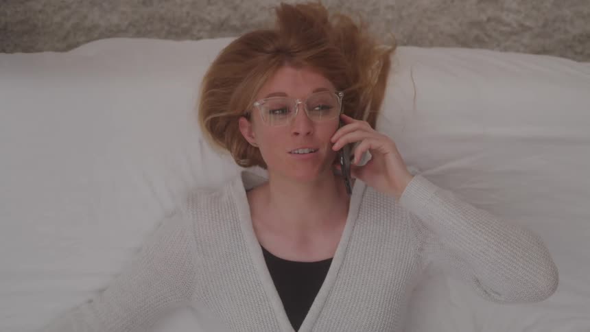 A woman with light brown hair and glasses lies on her back on a white bed talking on her cell phone