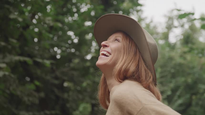 A woman wearing a hat laughs joyfully while looking up in a lush green outdoor setting
