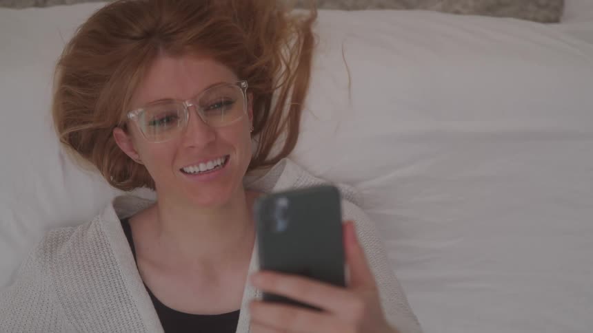 Happy woman laying down talking and waving while face timing on cell phone
