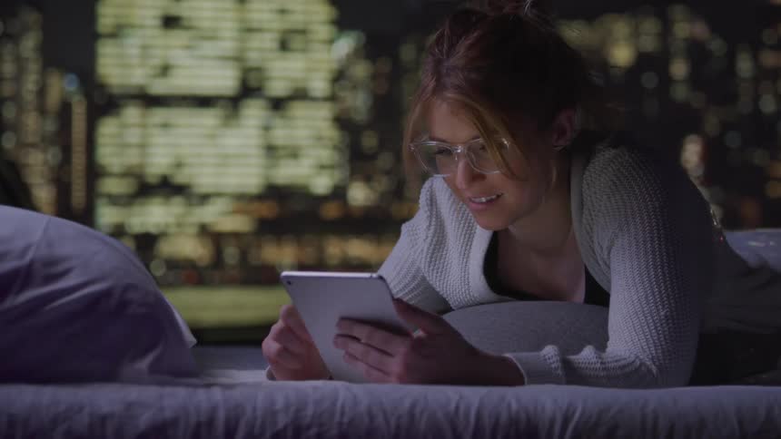 A woman with glasses lies in bed at night illuminated by the glow of a tablet with a blurred cityscape visible outside the window