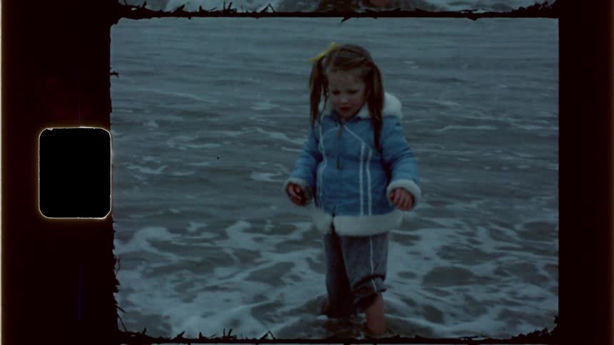 A little girl wearing winter clothes stands at the shoreline of an ocean on the beach