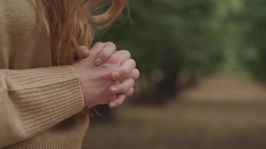 Womans hands clasped together her long light brown hair partially visible against a blurred natural background