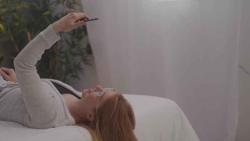 A woman with light brown hair lies on a white bed holding her phone up in a video call and waving