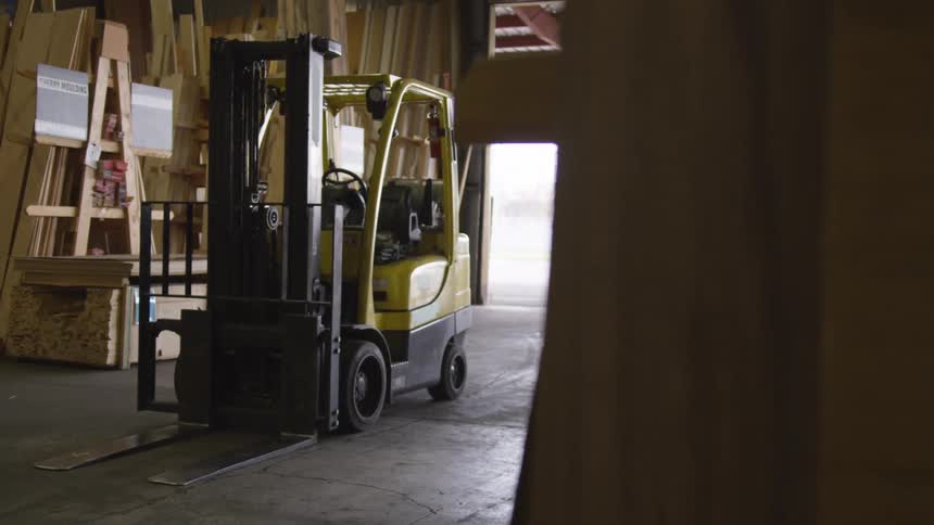 A yellow forklift is parked inside a woodshop surrounded by tall stacks of lumber and wooden planks