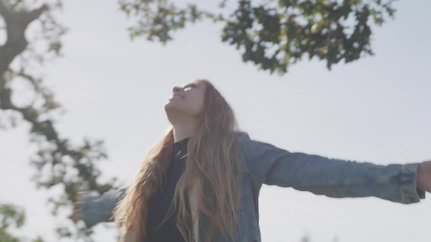 A woman with long light brown hair stands with her arms outstretched looking up towards the sky and a tree above her