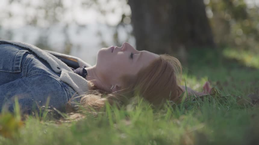 A woman with light brown hair lies on her back in the grass with her eyes closed wearing a denim jacket and with a tree trunk visible in the background