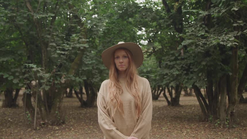 A woman with long light brown hair wearing a hat and a beige sweater stands facing the camera in a hazelnut orchard with tall leafy trees