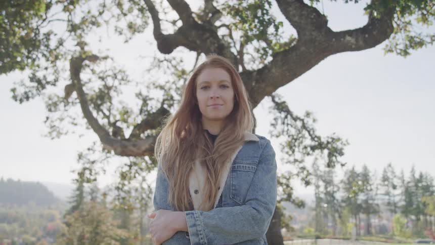 A woman with long reddish brown hair stands with her arms crossed looking directly at the camera with a large tree and a scenic landscape in the background
