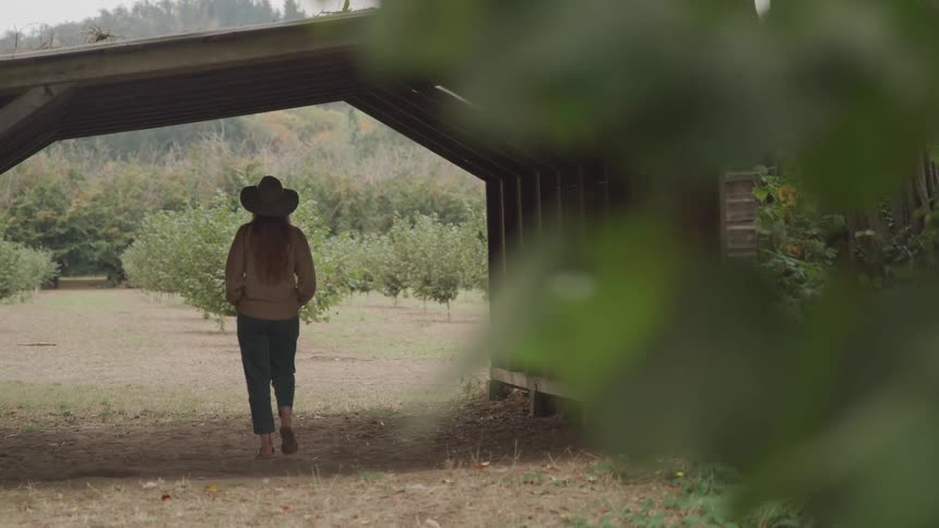 A woman wearing a hat is walking under a wood bridge on a path through the woods
