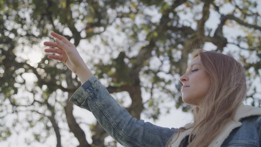 A woman with long light brown hair wearing a denim jacket reaches her arm up towards the sunlight filtering through tree branches with her eyes closed and face tilted upwards
