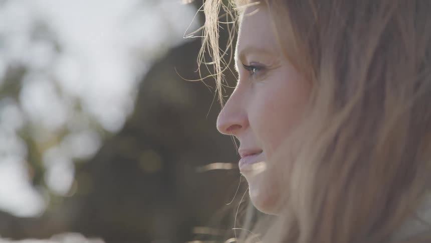 A woman with light colored hair her face partially illuminated by sunlight against a blurred background