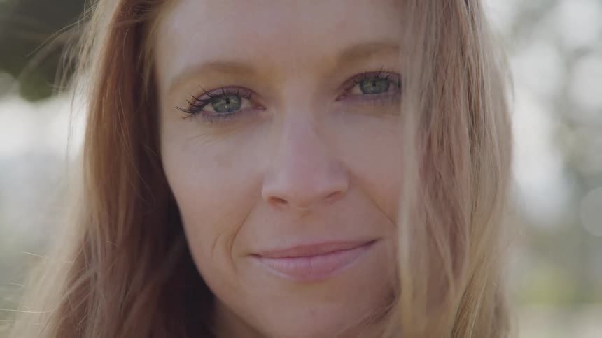 The face of a woman with light brown hair fair skin and blue eyes with a gentle smile and a slightly blurred background