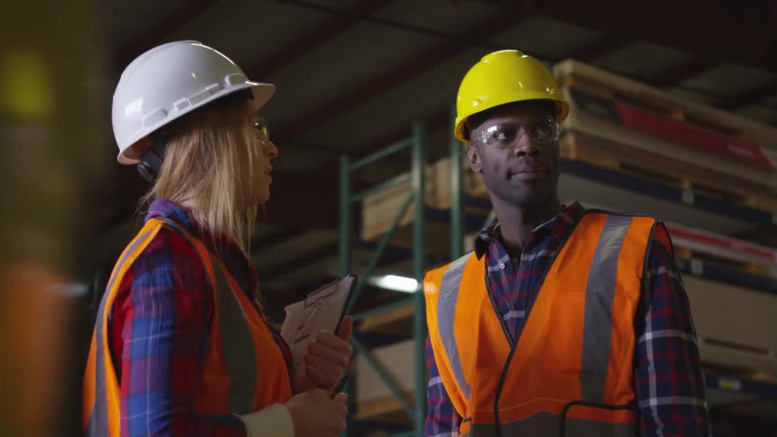 Two workers in hard hats and safety vests stand in a warehouse